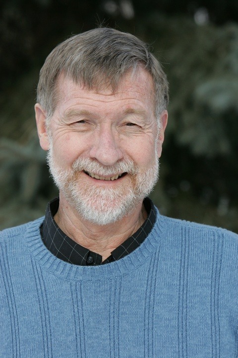 Smiling older man with a light blue sweater and a black collared shirt against a blurred green background, conveying warmth and approachability.